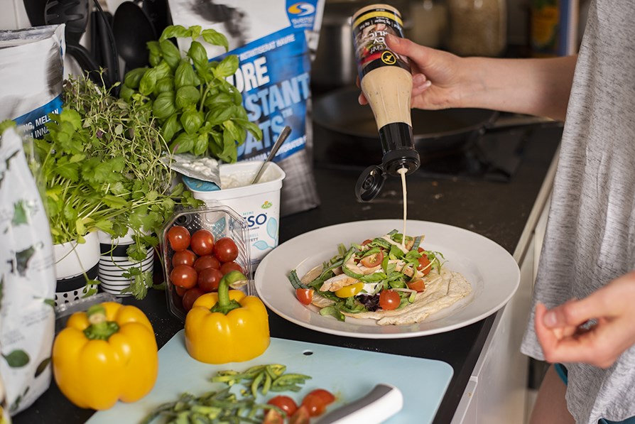 Henkil� lis�� kastiketta t�ytetyn wrapin p��lle. P�yd�ll� my�s Core Instant Oats, yrttej�, kirsikkatomaatteja, raejuustoa ja paprikaa.