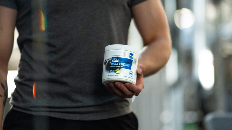 Henkil� seisoo sumean taustan edess� ja pitelee Core BCAA Energy -purkkia k�dess��n.
