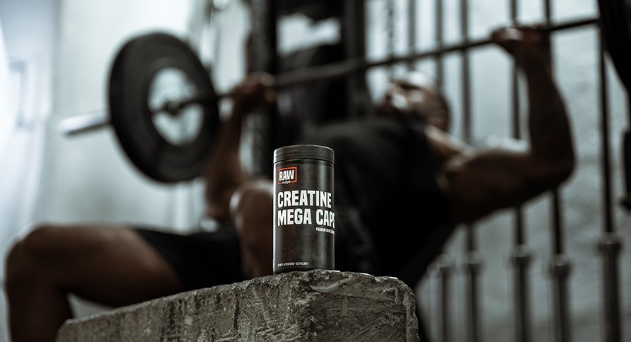 RAW Creatine Mega Caps keski�ss�, taustalla mies suorittamassa penkkipunnerrusta vinopenkiss�.
