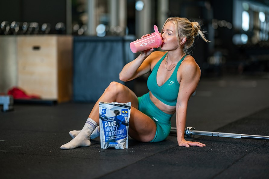 Nainen istuu lattialla kuntosalilla ja juo Core Whey Proteiinia shakerista. 