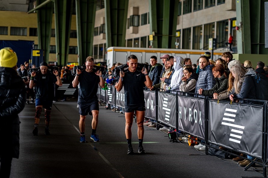 Hyrox-kilpailu, jossa miehet tekevt askelkyykkyj hiekkaskin kanssa.