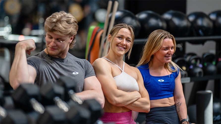 Tv� kvinnor och en man st�r bredvid varandra i gymmilj� ikl�dda tr�ningskl�der. Kvinnan i mitten har armarna i kors och ler mot kameran.
