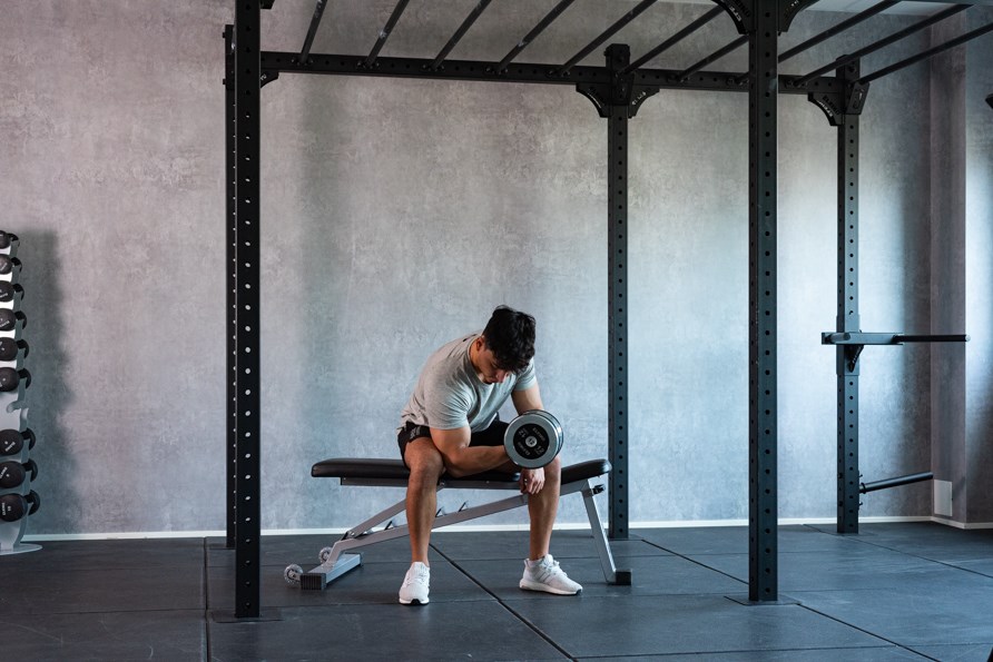 Tummahiuksinen mies treenaa hauiskääntöä vinopenkillä istuen. Tummahiuksinen mies treenaa hauiskääntöä vinopenkillä istuen.