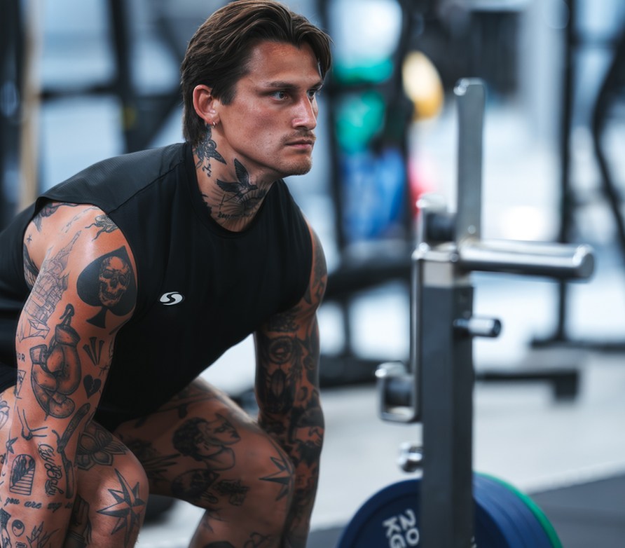 Tatuoitu tummahiuksinen mies maastavedon lähtöasennossa keskittynyt ilme kasvoillaan. Tatuoitu tummahiuksinen mies maastavedon lähtöasennossa keskittynyt ilme kasvoillaan.