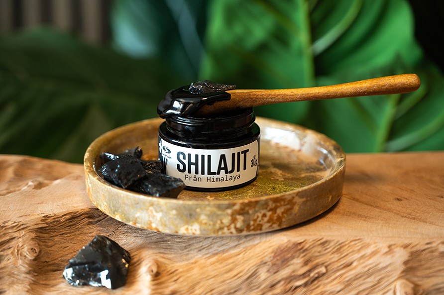 Avattu Healthwell Shilajit -purkki pienen keraamisen lautasen p��ll� puisella alustalla. Taustalla vihreit� lehti�.